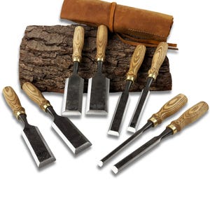 Puede incluir: Un juego de ocho cinceles de madera con mangos de madera y hojas de metal. Los cinceles están dispuestos sobre un trozo de corteza, con un rollo de herramientas de cuero marrón en el fondo. Los cinceles varían en tamaño y forma de hoja.