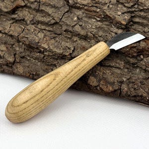 May include: A wood carving knife with a light-colored wooden handle and a silver blade. The blade is angled and appears sharp. The handle is smooth and rounded, designed for a comfortable grip. The knife is designed for wood carving.