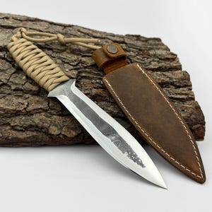 May include: A handmade dagger with a tan wrapped handle and a polished silver blade. The dagger is paired with a brown leather sheath. The knife is resting on a piece of textured wood.