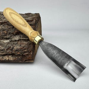 以下が含まれることがあります： 明るい色の木製ハンドルと真鍮のフェルールが付いた木彫りのガウジ。 刃は濃い色の金属でできており、湾曲した形状をしています。 このツールは木工用に設計されており、質感のある木材の上に置かれています。