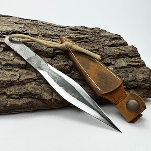 May include: A hand-forged, silver-colored throwing knife with a sharp point, lying on a piece of textured wood. A brown leather sheath with a button closure and a tan cord is next to the knife. The knife is a handcrafted item.