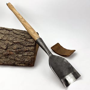 May include: A hand-forged, heavy-duty wood carving gouge with a light-colored wooden handle and a dark metal blade. A small, brown leather sheath rests nearby. The gouge is angled against a piece of textured wood.