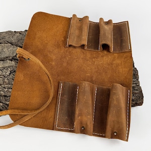 May include: A brown leather tool roll with multiple pockets and a leather tie closure. The roll is made of distressed leather and features white stitching. The tool roll is designed to hold various tools and accessories.