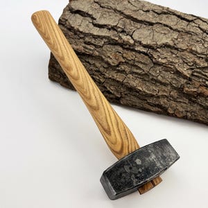 May include: A heavy-duty sledgehammer with a light brown wooden handle and a dark gray head. The handle has a textured wood grain pattern. A piece of tree bark rests in the background.
