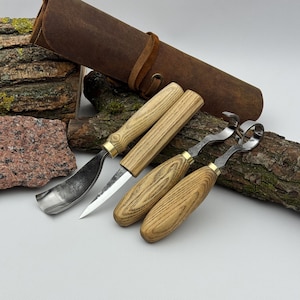 May include: A set of four woodworking carving tools with light-colored wooden handles and brass ferrules, including a hook knife, a detail knife, and two gouges, accompanied by a brown leather roll-up storage case with a leather tie closure.