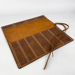 May include: Open, brown leather roll-up case with white stitching. The case has multiple compartments and a leather tie closure. The interior is a lighter shade of brown than the exterior. The case is designed to hold items such as pens or tools.