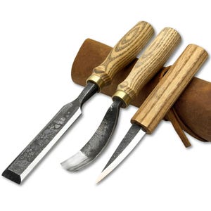 May include: A set of three wood carving tools with wooden handles and metal blades. The tools include a chisel, a curved gouge, and a knife, all resting in a brown leather sheath. The handles have a light wood grain pattern.