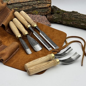 May include: A set of woodworking chisels and gouges with wooden handles and metal blades, displayed on a brown leather tool roll. The chisels have brass ferrules. The background includes wood and stone.