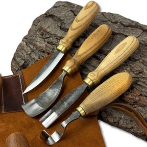 May include: A set of four wood carving tools with wooden handles and metal blades. The blades have different shapes, including a straight blade, a curved blade, and a gouge. The tools are arranged on a brown leather surface.