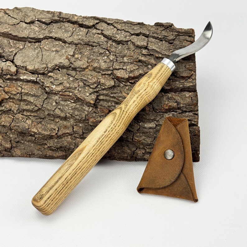 May include: A wood carving knife with a curved blade and a wooden handle. The knife is resting on a piece of bark. A brown leather sheath is included.