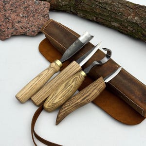 Könnte beinhalten: Ein Set aus vier Holzschnitzwerkzeugen mit hellen Holzgriffen und Metallklingen, bestehend aus einem Hohleisen, einem geraden Messer, einem Hakenmesser und einem Detailmesser, angeordnet auf einer braunen Leder-Werkzeugrolle.