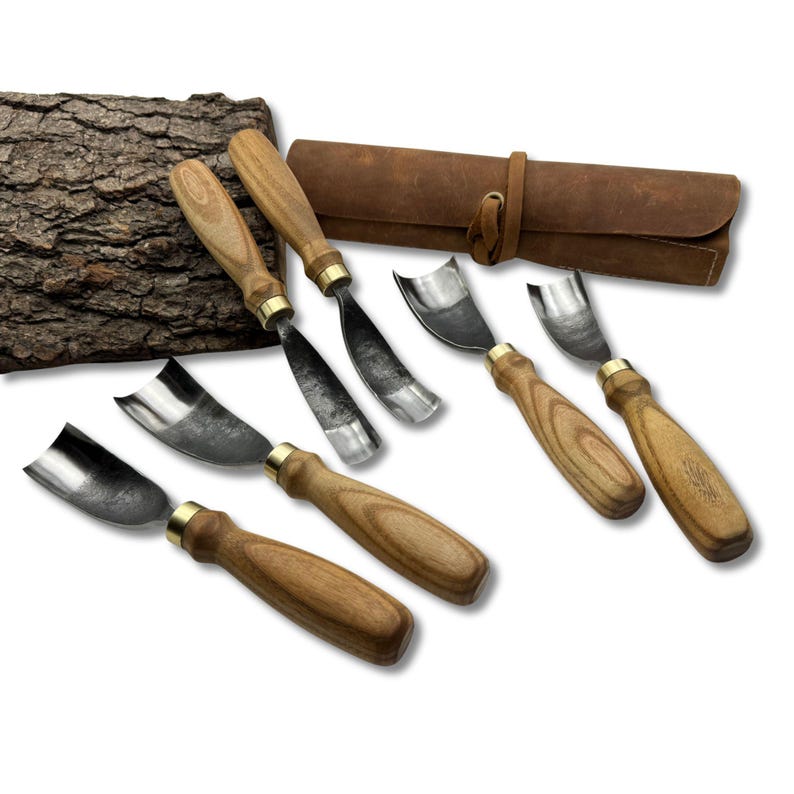 May include: A set of wood carving tools with wooden handles and metal blades. The tools are arranged with a leather roll-up case and a piece of bark in the background. The handles have a brass ferrule.