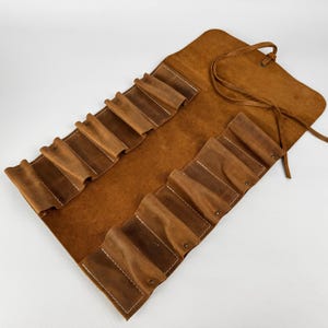 Könnte beinhalten: Ein braunes Leder-Stifteetui zum Aufrollen mit mehreren Taschen. Das Etui besteht aus einem Leder mit Used-Look und verfügt über weiße Nähte und einen Lederverschluss. Die Taschen sind für Stifte, Bleistifte oder andere kleine Kunstutensilien konzipiert.