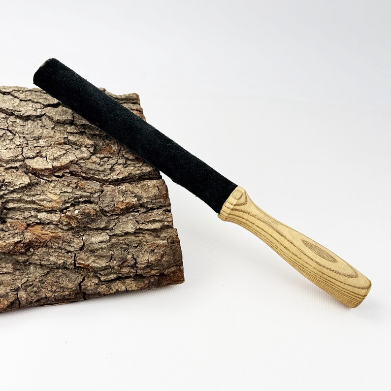 May include: A singing bowl mallet featuring a long, cylindrical black felt-covered head attached to a smooth, light-colored wooden handle with a visible grain pattern. The mallet is positioned diagonally against a piece of rough tree bark.