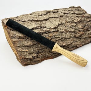 May include: A long, cylindrical tool featuring a black, textured, felt-like covering on the upper portion and a light-colored, natural wood handle with a visible grain pattern at the base, designed for manual use and grip.
