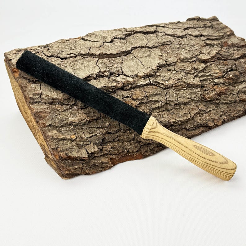 May include: A long, cylindrical tool featuring a black, textured, felt-like covering on the upper portion and a light-colored, natural wood handle with a visible grain pattern at the base, designed for manual use and grip.