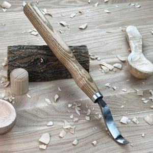 May include: A wooden carving tool with a curved blade and a wooden handle. The tool is lying on a wooden surface with wood shavings scattered around it. There is also a partially carved wooden spoon in the background.