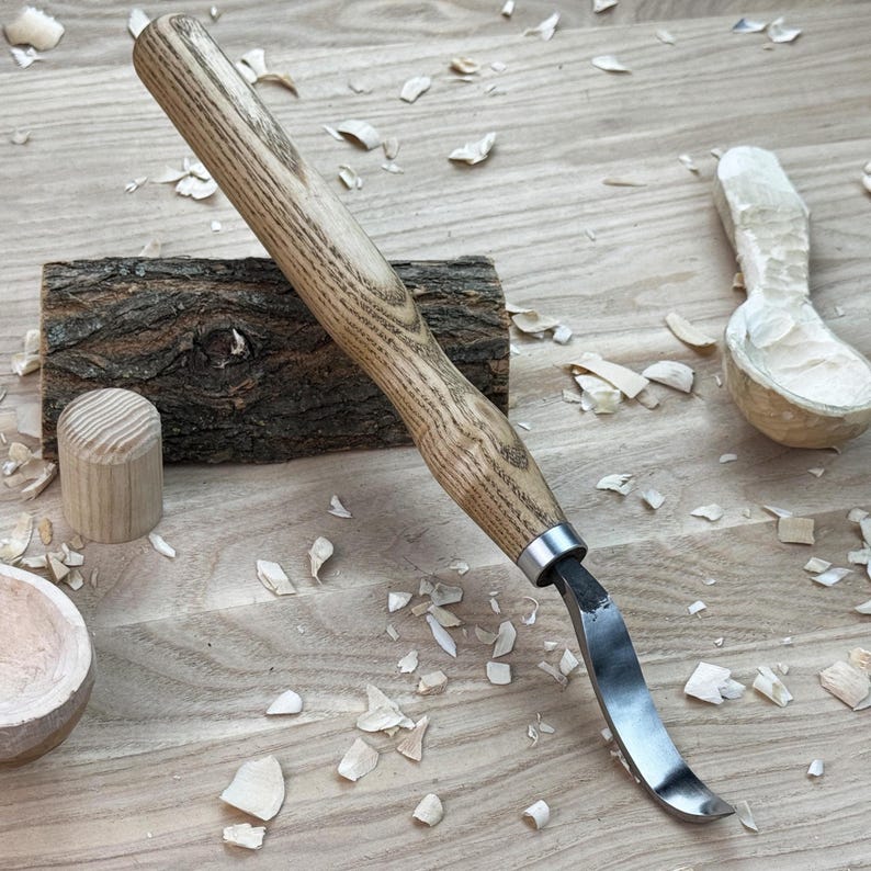 May include: A wooden carving tool with a curved blade and a wooden handle. The tool is lying on a wooden surface with wood shavings scattered around it. There is also a partially carved wooden spoon in the background.