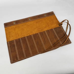 May include: Open leather roll-up case in shades of brown and tan. The case has multiple vertical pockets with white stitching. A leather tie secures the case when rolled. The interior is a lighter tan colour.