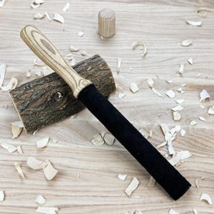 May include: A long, cylindrical tool featuring a light-colored, wood-grained handle and a solid black, textured shaft. The tool rests diagonally across a rough, bark-covered wooden log, with a small, separate wooden cylinder nearby.