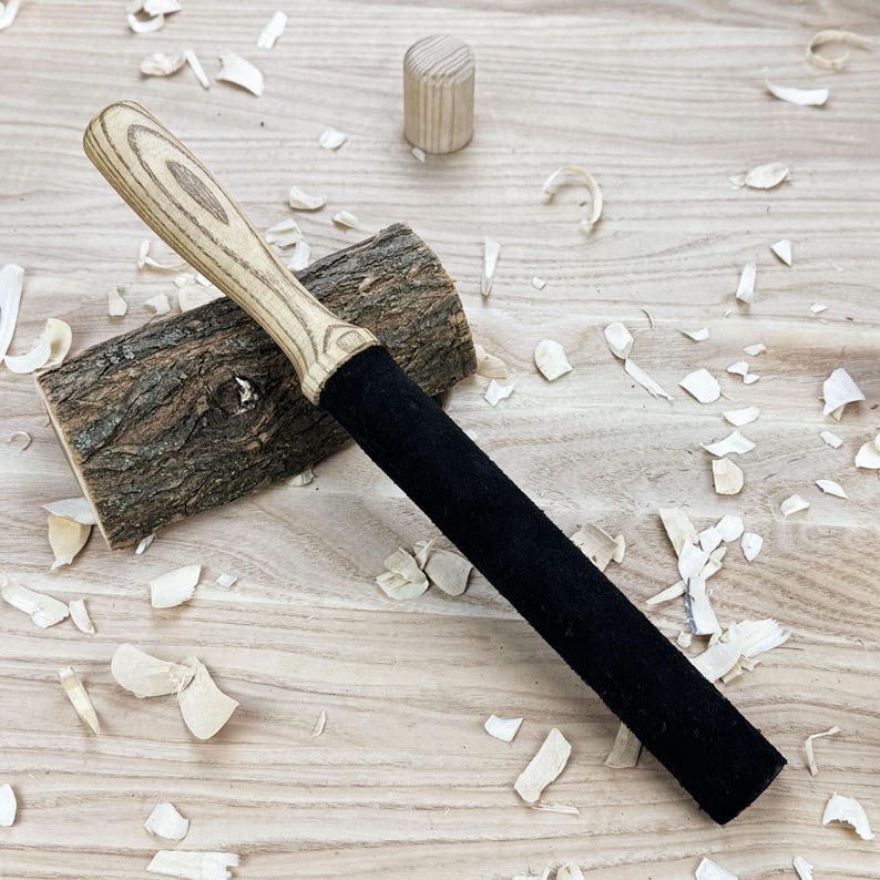May include: A long, cylindrical tool featuring a light-colored, wood-grained handle and a solid black, textured shaft. The tool rests diagonally across a rough, bark-covered wooden log, with a small, separate wooden cylinder nearby.