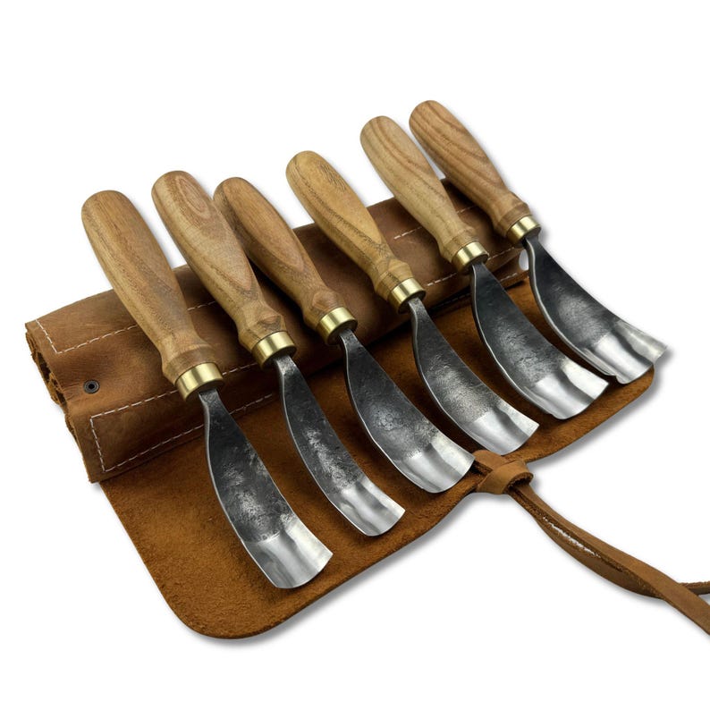 May include: A set of six wood carving chisels with wooden handles and metal blades, presented in a brown leather tool roll. The chisels have a curved blade design and are arranged in a row. The tool roll is open, revealing the tools.