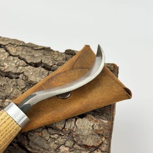 May include: A curved wood carving knife with a brown leather sheath. The knife has a wooden handle and a metal blade.