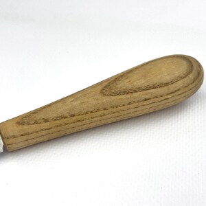 May include: Close-up of a wooden handle for a tool or utensil. The handle is made of light brown wood with a grain pattern. The handle is oval-shaped and has a smooth finish.