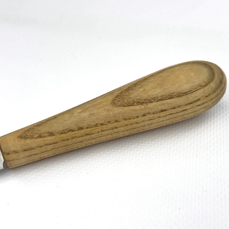 May include: Close-up of a wooden handle for a tool or utensil. The handle is made of light brown wood with a grain pattern. The handle is oval-shaped and has a smooth finish.