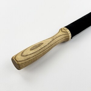 May include: A tool featuring a light-colored wooden handle with a prominent grain pattern and a smooth, contoured shape. The handle is attached to a long, cylindrical section covered in a solid black, textured material that extends outward.