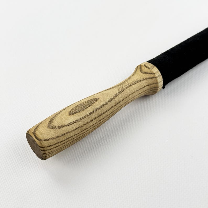 May include: A tool featuring a light-colored wooden handle with a prominent grain pattern and a smooth, contoured shape. The handle is attached to a long, cylindrical section covered in a solid black, textured material that extends outward.