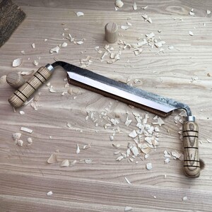 以下が含まれることがあります： 革製の鞘と木製のハンドルが付いた湾曲した木彫りナイフ。ナイフは木製の表面に置かれ、その周りに木くずが散らばっています。