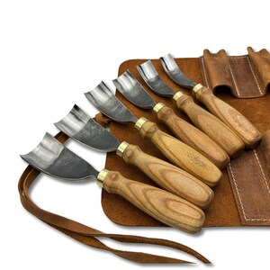 May include: A set of wood carving tools with curved blades and wooden handles. The tools are displayed on a brown leather roll-up case with a strap. The blades are silver, and the handles are light brown with brass ferrules.