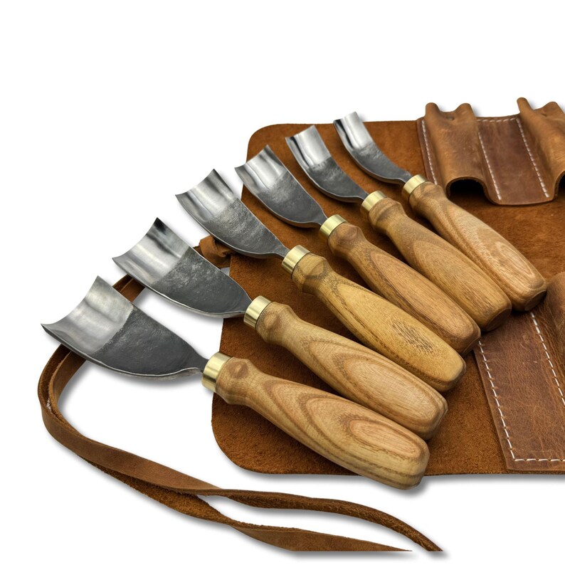 May include: A set of wood carving tools with curved blades and wooden handles. The tools are displayed on a brown leather roll-up case with a strap. The blades are silver, and the handles are light brown with brass ferrules.
