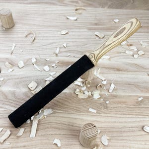 May include: A leather stropping tool featuring a long, cylindrical black leather surface attached to a light-colored, grained wooden handle. The handle displays a distinct wood grain pattern, and the tool is designed for sharpening blades.