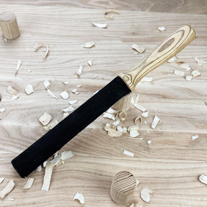 May include: A leather stropping tool featuring a long, cylindrical black leather surface attached to a light-colored, grained wooden handle. The handle displays a distinct wood grain pattern, and the tool is designed for sharpening blades.