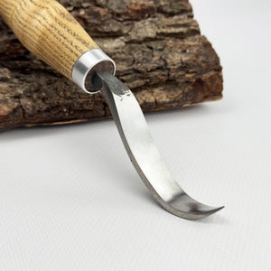May include: A curved wood carving knife with a wooden handle and a metal ferrule. The blade is curved and has a sharp point.