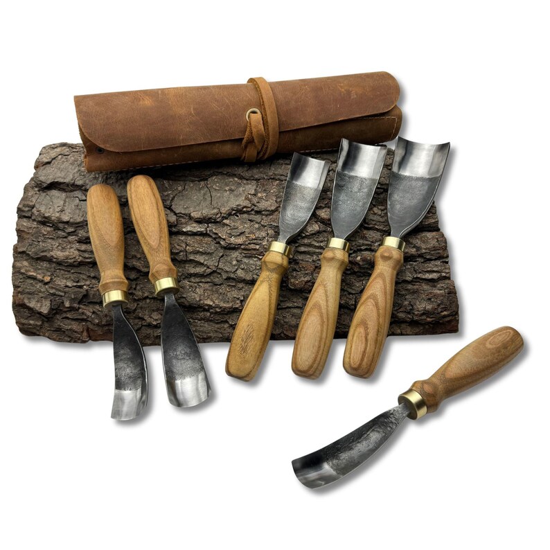 May include: A set of wood carving tools with wooden handles and metal blades, arranged on a piece of bark. A brown leather tool roll is in the background. The tools have brass accents.
