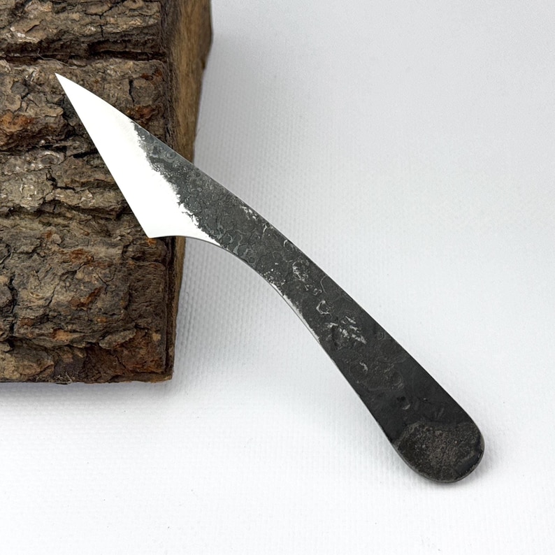 May include: A small, hand-forged knife with a curved, textured handle and a sharp, silver blade. The blade has a pointed tip and a rough, unfinished edge. The knife is set against a piece of wood with a textured bark.