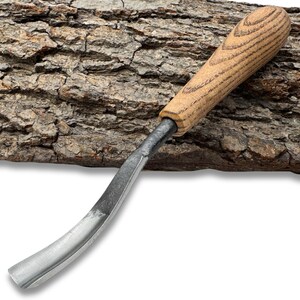Può includere: Uno scalpello per intaglio del legno con una lama curva color argento e un manico in legno marrone chiaro. L'impugnatura presenta un motivo a venature strutturate. Lo strumento è progettato per modellare legno e altri materiali.