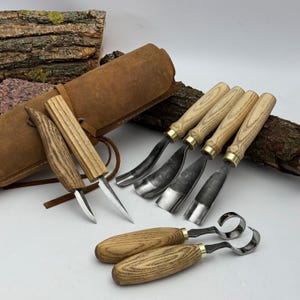 May include: A set of wood carving tools with wooden handles and metal blades. The tools are arranged with a brown leather roll-up case and a piece of wood in the background. The tools include gouges, knives, and hook knives.
