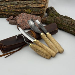 May include: A set of three wood carving tools featuring steel blades and light-colored wooden handles with brass ferrules. The set includes a hook knife, a straight blade knife, and a gouge, all resting on a dark brown leather tool roll.