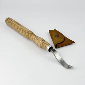 May include: A wooden handled carving tool with a curved blade and a leather sheath. The tool is made of wood and metal and has a brown leather sheath.