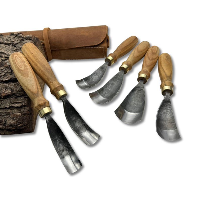 May include: A collection of six wood carving gouges, each with a wooden handle and a metal blade. The tools are displayed with a brown leather tool roll and a piece of bark. The gouges feature a curved blade design.
