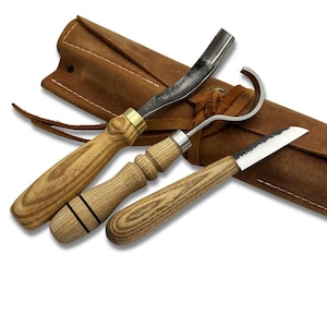 May include: A set of three wood carving tools with wooden handles and metal blades, alongside a brown leather carrying case. The tools include a curved gouge, a hook knife, and a straight knife. The leather case has a drawstring closure.