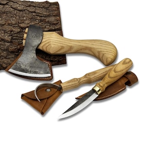 Puede incluir: Un hacha pequeña, un cuchillo de gancho y un cuchillo para tallar con mangos de madera y fundas de cuero. El hacha tiene una cabeza de metal y un mango de madera curvado. El cuchillo de gancho tiene una hoja curva.