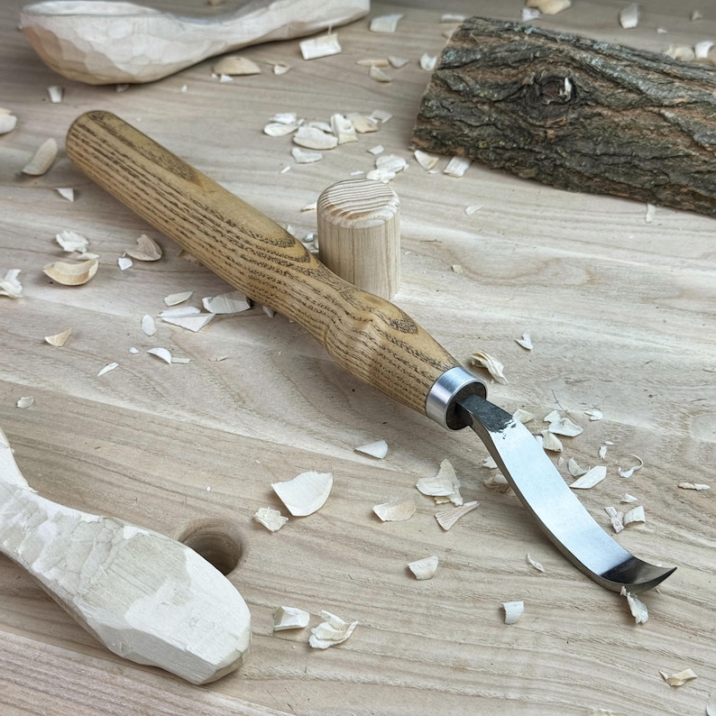 May include: A wooden carving tool with a curved blade and a wooden handle. The tool is lying on a wooden surface with wood shavings around it.