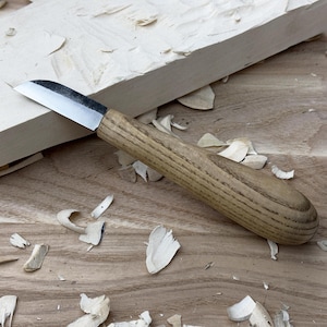 May include: A close-up of a wood carving knife with a wooden handle. The knife is lying on a wooden surface with wood shavings scattered around it.