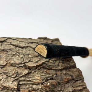 May include: A cylindrical object with a black suede-like exterior and a semi-circular wooden end cap. The object features a smooth, dark fabric texture along its length, contrasting with the light brown, natural wood grain visible on the flat, semi-circular cross-section at the tip.