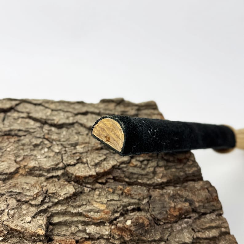 May include: A cylindrical object with a black suede-like exterior and a semi-circular wooden end cap. The object features a smooth, dark fabric texture along its length, contrasting with the light brown, natural wood grain visible on the flat, semi-circular cross-section at the tip.